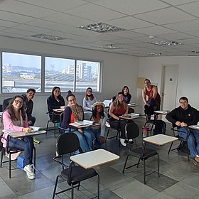 Eine Gruppe von jungen Leuten sitzt in einem Klassenraum an Einzeltischen. Sie lächeln in die Kamera. Im Hintergrund kann man durch das Fenster die Skyline von São Paulo erkennen. 