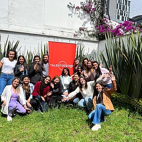 Un grupo de estudiantes de idiomas en el jardín de la escuela de idiomas TalentOrange de Bogotá. Están sentados en un prado. Al fondo hay una pared blanca y un arbusto con flores moradas. Todos ríen alegremente.